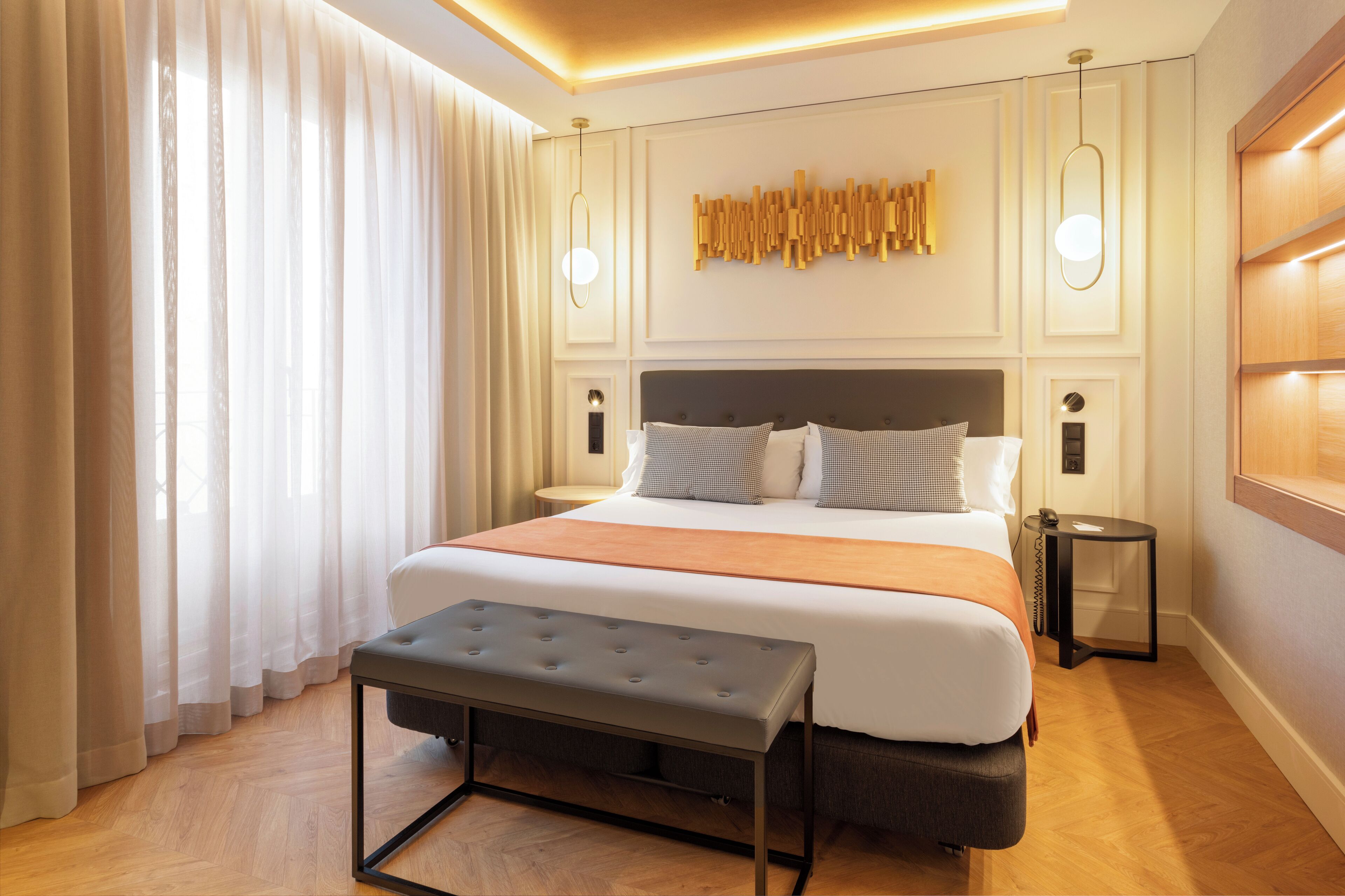 A modern hotel room with a large bed, a bench, a bedside table, and a wall-mounted light fixture.