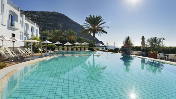 Ein klares, blaues Schwimmbecken mit Fliesenboden, Liegestühlen, Sonnenschirmen und einem Berg im Hintergrund.
