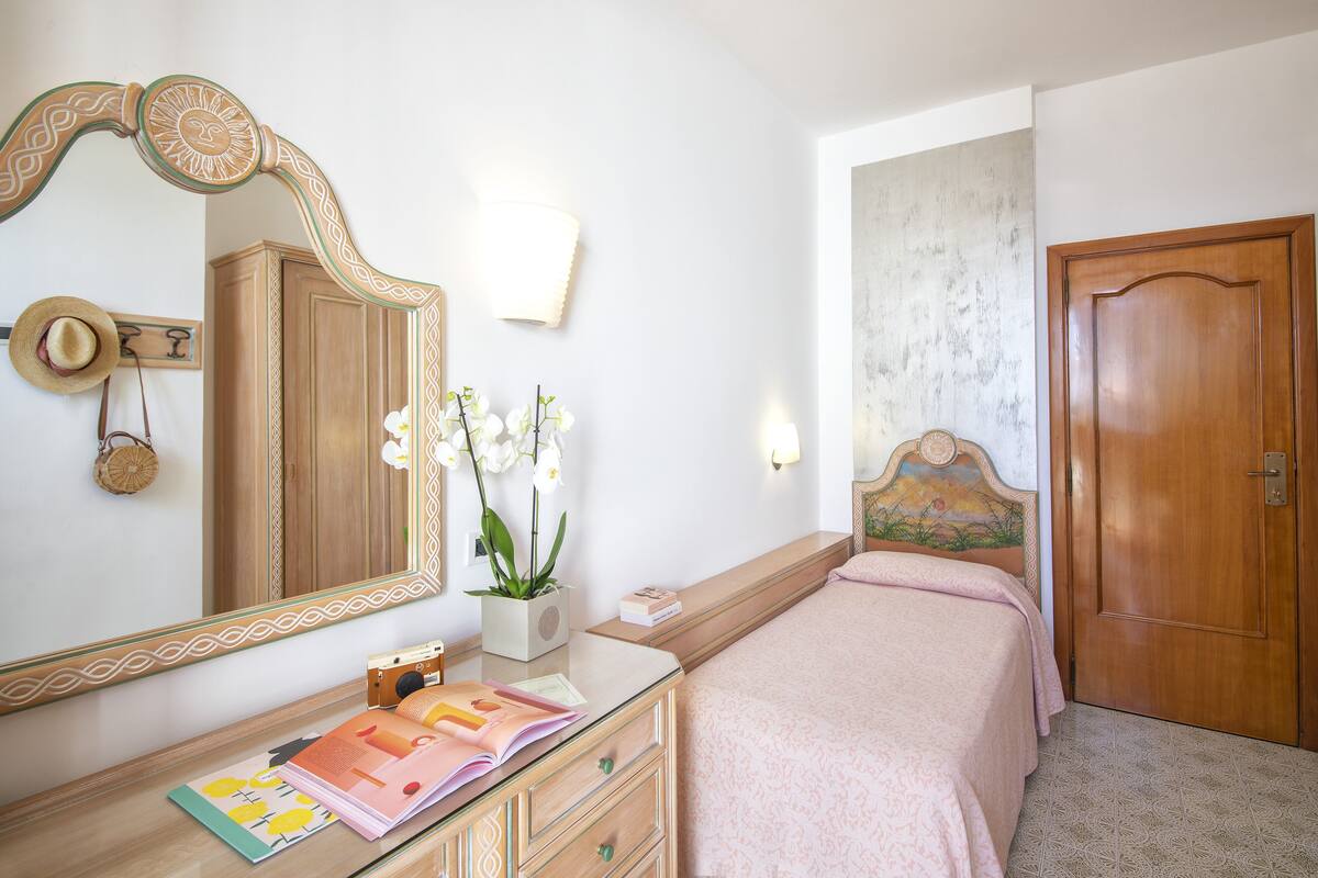 Un dormitorio con una puerta de madera, un espejo grande con marco decorativo, una mesita de noche con lámpara y una cama con colcha rosa.