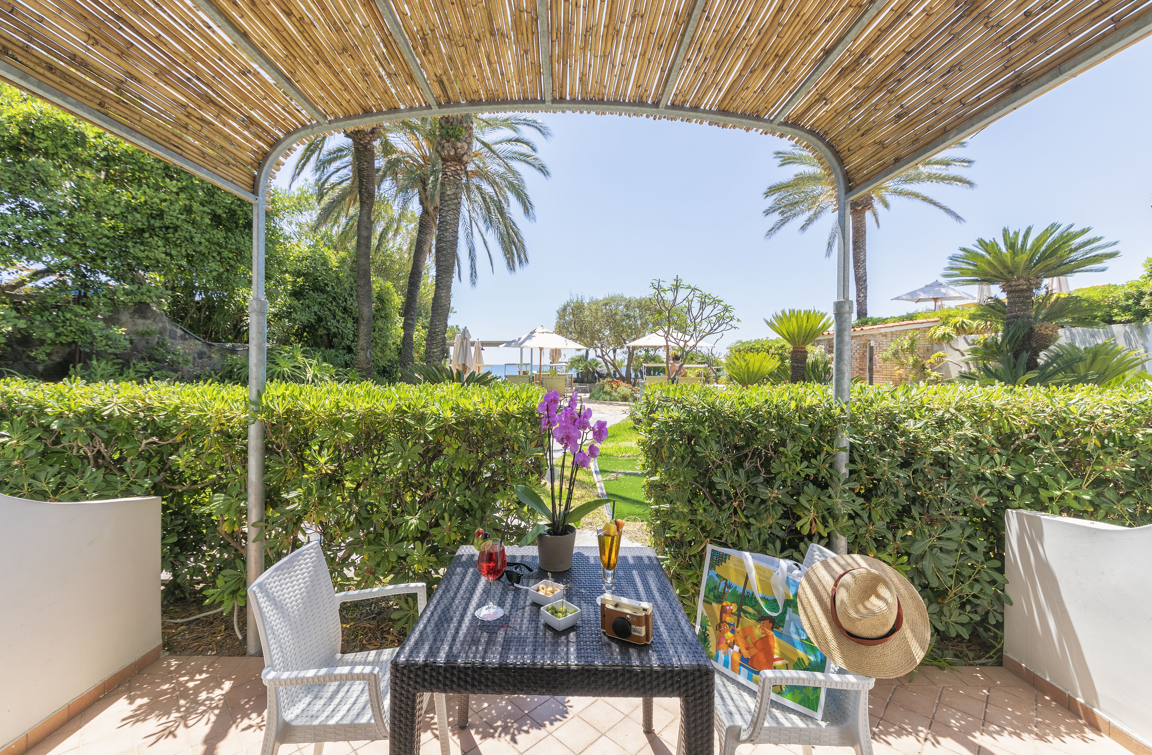 An outdoor patio with a table set for two, surrounded by greenery and palm trees.