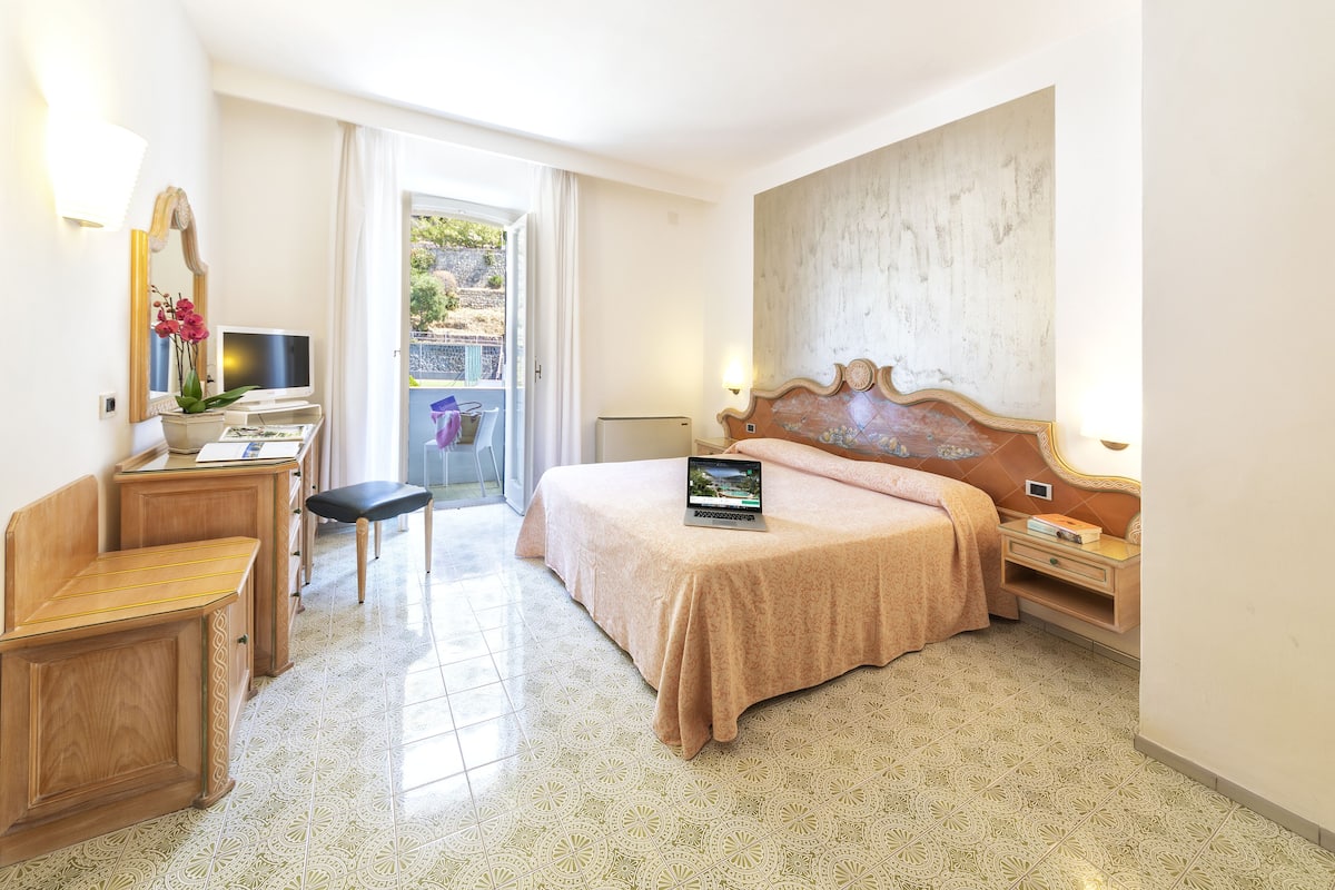 Una habitación de hotel con cama, escritorio con una computadora portátil, silla, una mesita y ventana con vistas al exterior.