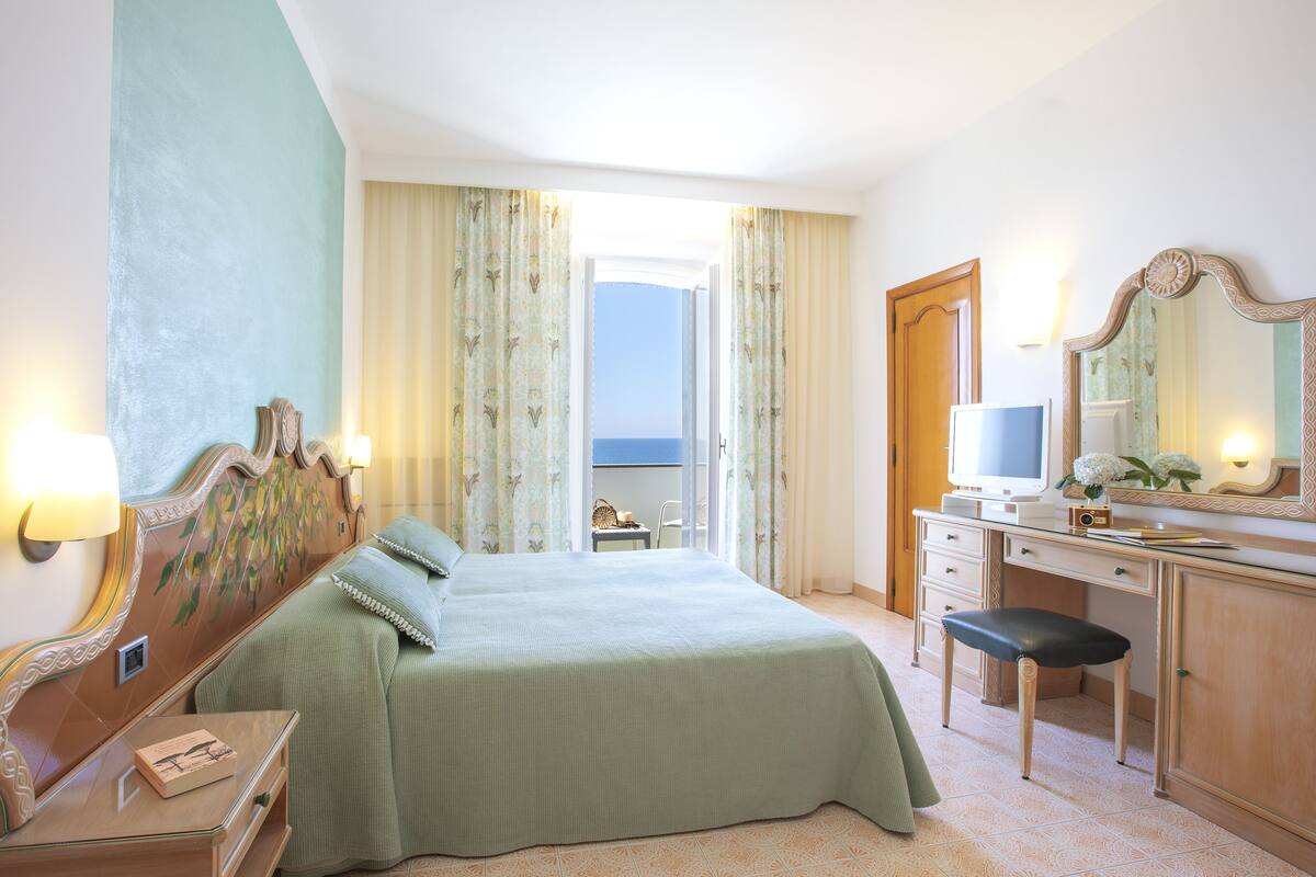 Un cuarto de hotel con cama, escritorio, silla, cómoda, espejo y vista al mar a través de la ventana.