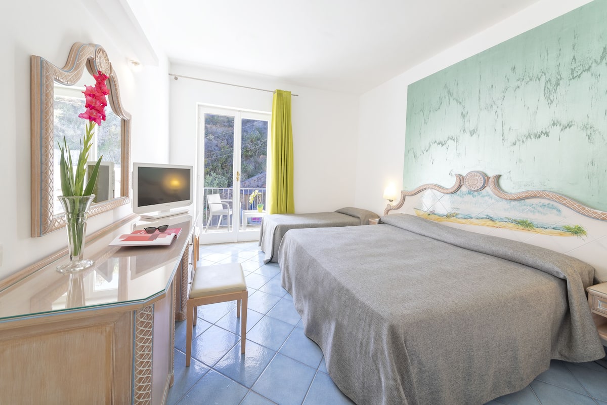 Hotellihuone, jossa on kaksi sänkyä, työpöytä tietokoneella, maljakko kukkineen ja suuri taulu seinällä.