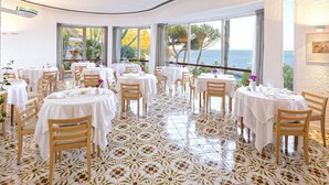 Un restaurante con mesas y sillas redondas, ventanas amplias con vistas al mar y un suelo de baldosas con un diseño floral.