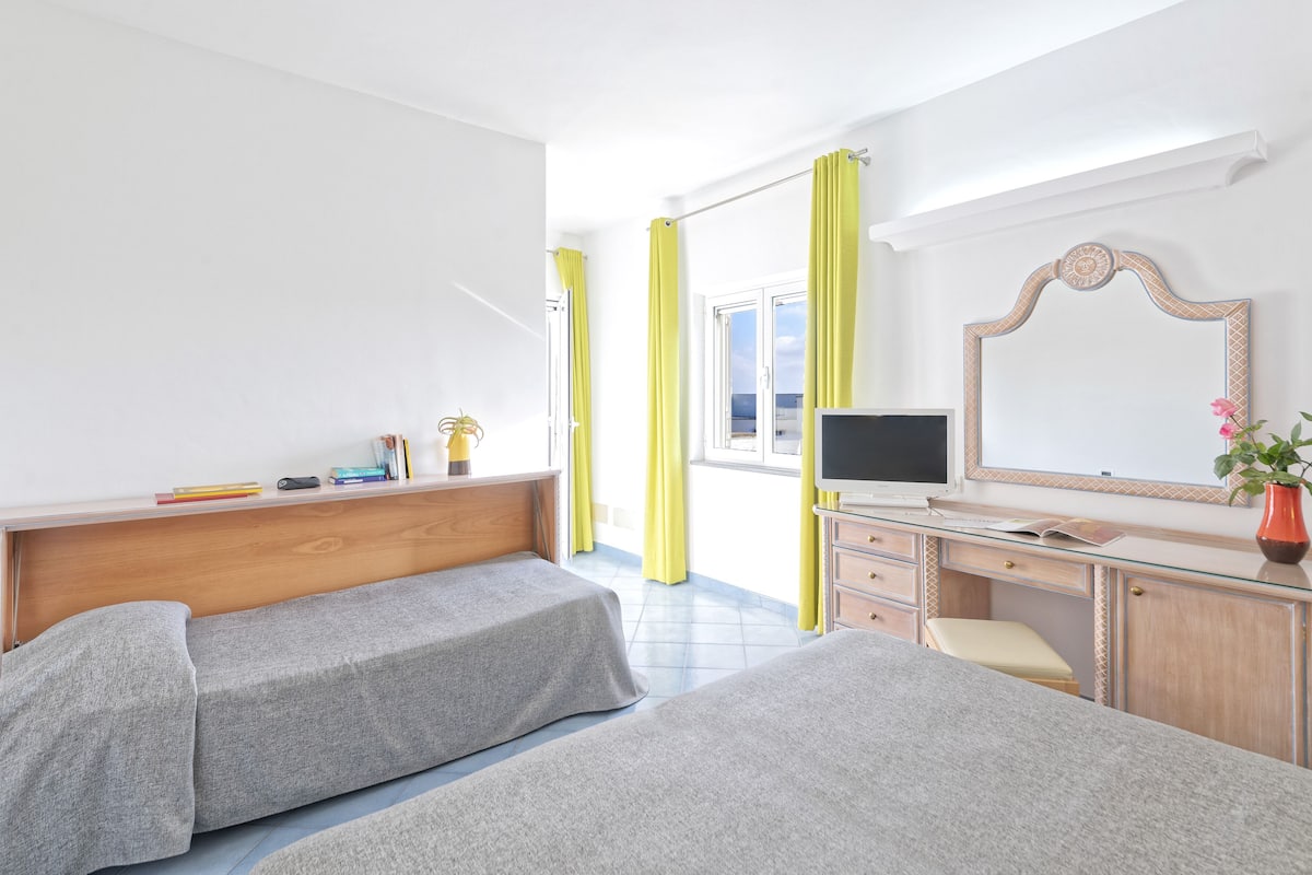 Hotellihuone, jossa on kaksi sänkyä, työpöytä tietokoneella, peili ja ikkuna, jossa on keltaiset verhot.