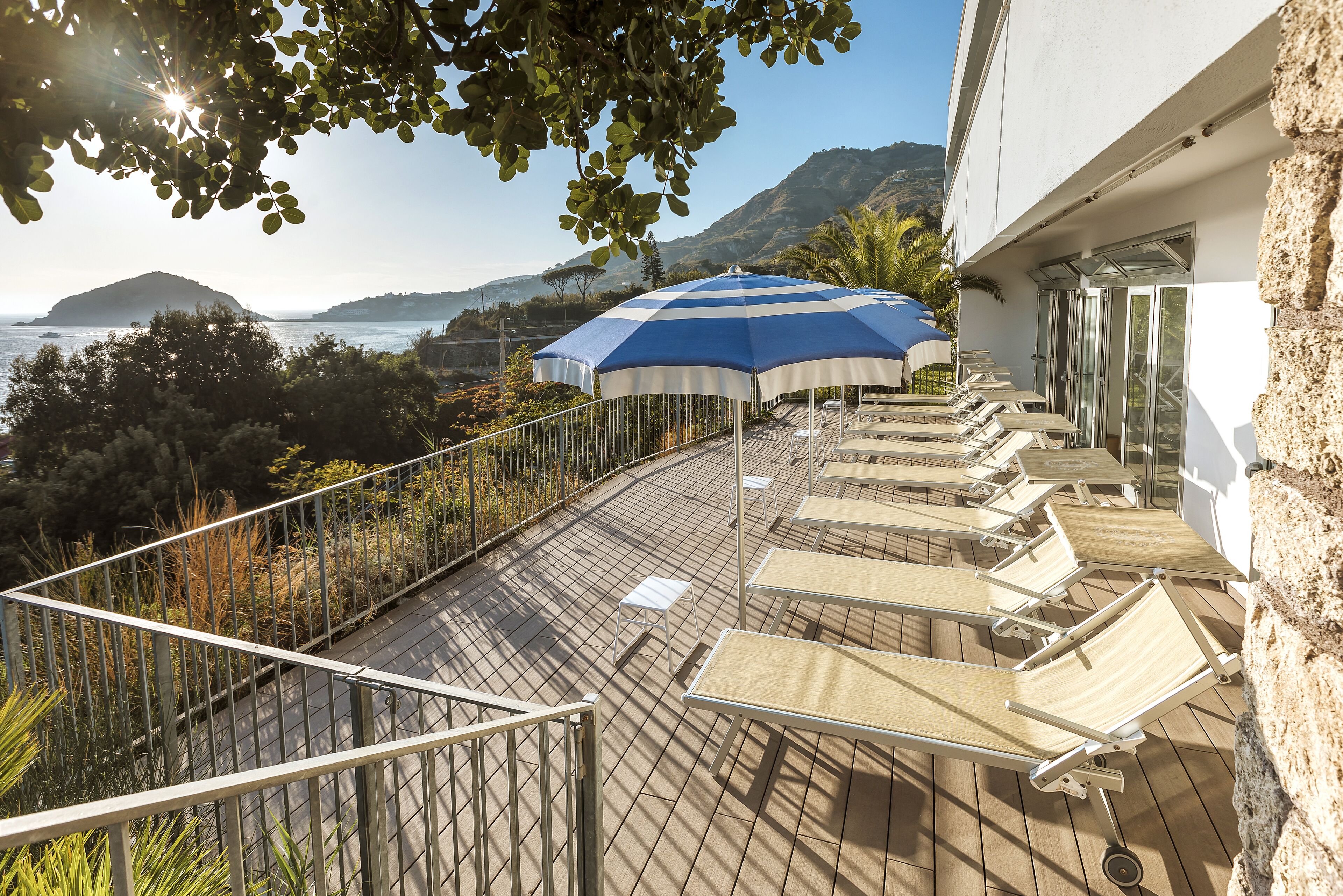 A terrace with sun loungers, a blue and white umbrella, and a view of the sea and hills.