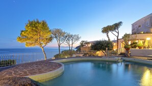 Zona de piscina con vista al mar, rodeada de árboles y edificios.