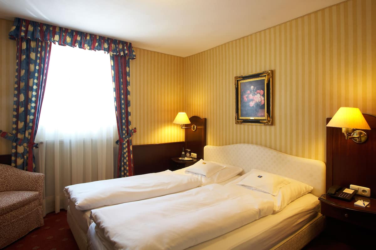 Habitación de hotel con dos camas, una silla, una mesita de noche con lámpara y un cuadro en la pared.