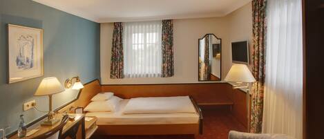 A hotel room with a bed, a desk with a lamp, a chair, a TV, and a window with curtains.