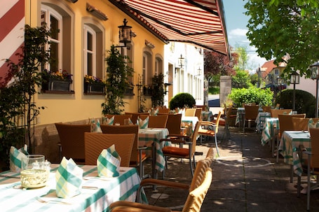 An outdoor cafe with wicker chairs and tables, covered by a striped awning, and surrounded by plants and buildings.