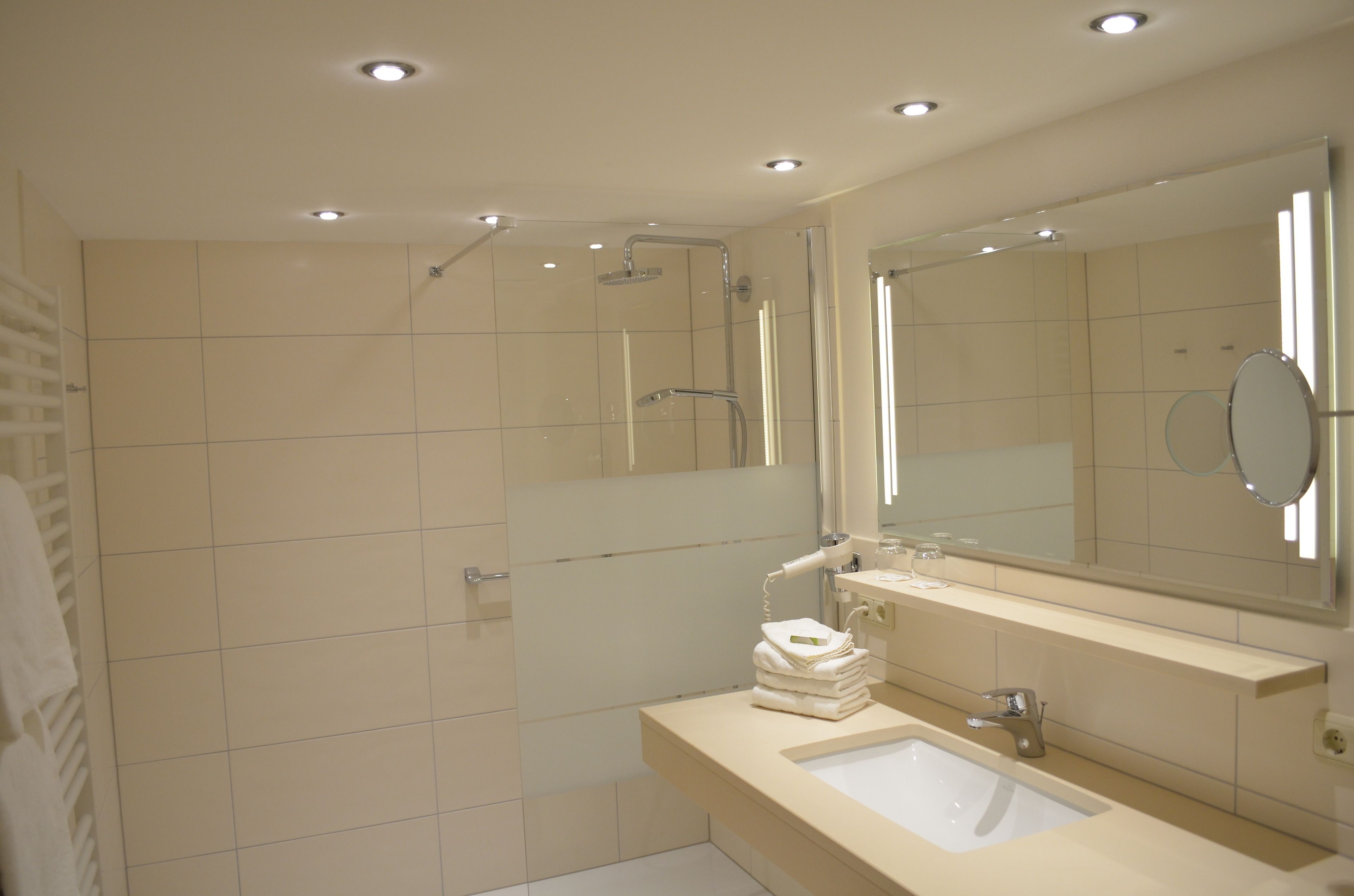 Une salle de bain moderne avec une douche à parois de verre, un grand miroir et un lavabo surmonté d’une pile d’essuie-tout.