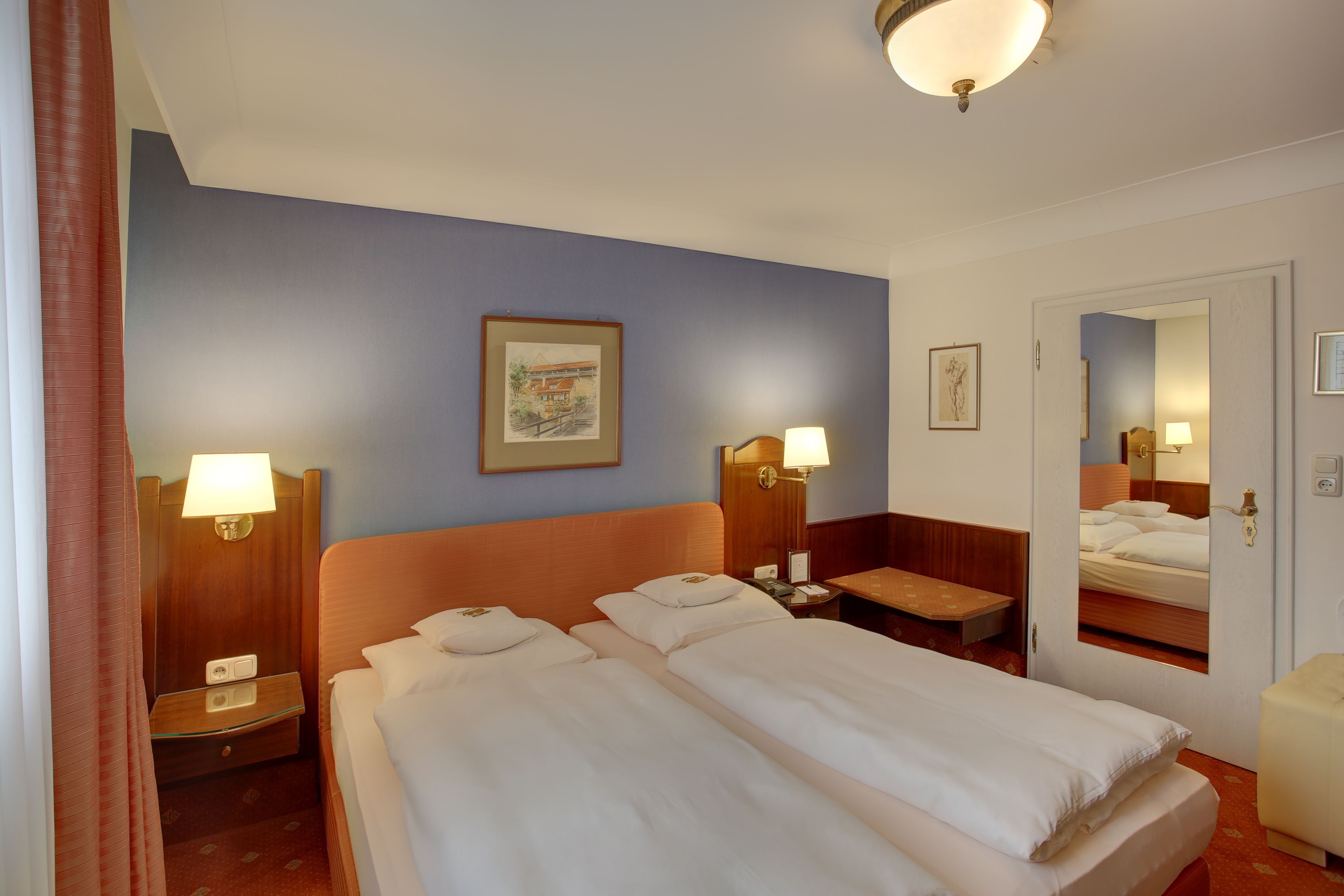 Habitación de hotel con dos camas, una mesita de noche con lámpara, una pequeña mesa y un espejo en la pared.