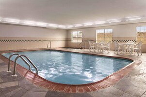 Una piscina rectangular techada con borde de azulejos, rodeada por un deck de piedra. Hay sillas de plástico blanco y una mesa cerca de una ventana con vistas.