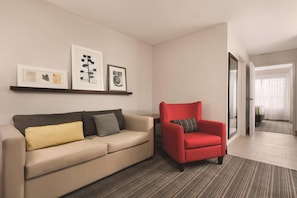 A living room with a beige sofa, a red armchair, and a coffee table. There are framed pictures on the wall and a door leading to another room.