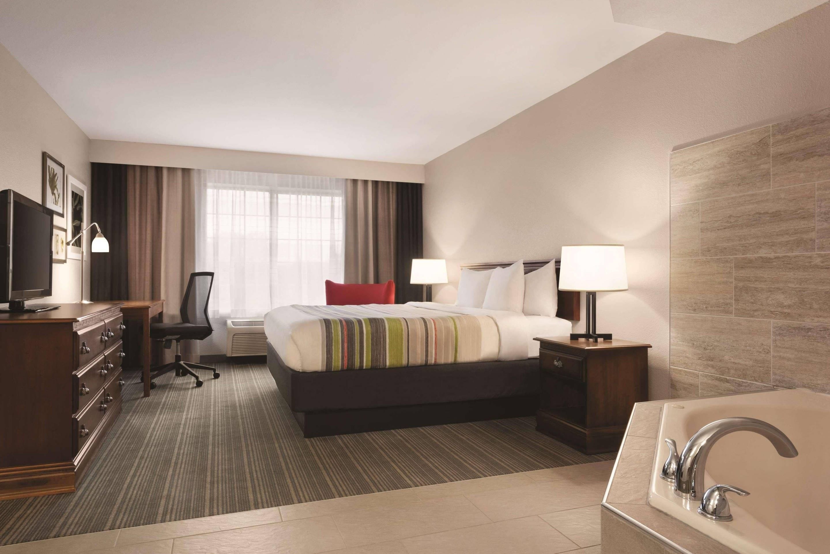 A hotel room with a bed, a desk, a chair, a TV, a lamp, a bathroom with a sink and a bathtub.