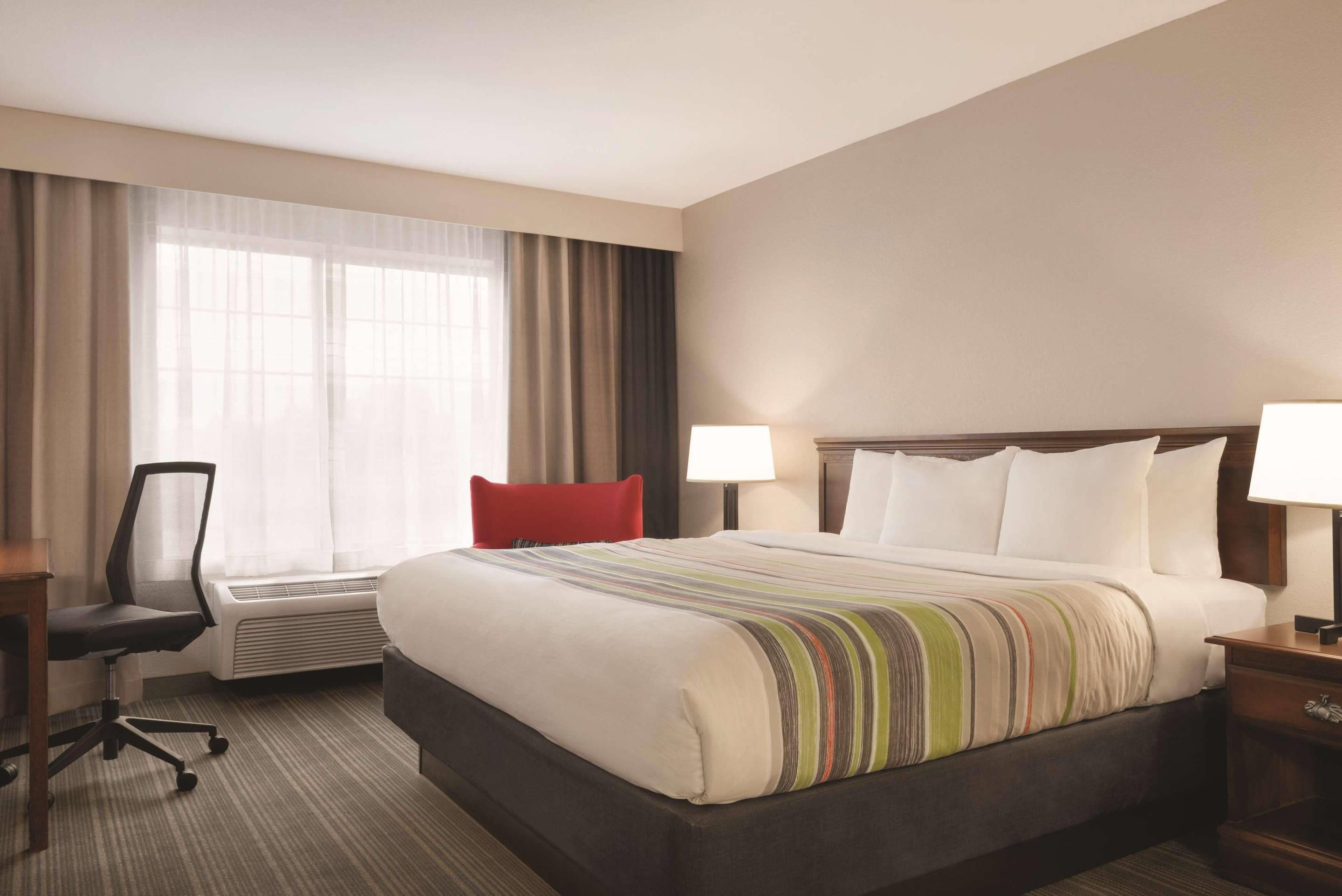 A hotel room with a large bed, a desk with a chair, a window with curtains, and a bedside table with a lamp.