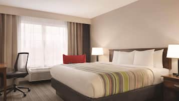 A hotel room with a large bed, a desk with a chair, a window with curtains, and a bedside table with a lamp.