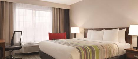 A hotel room with a large bed, a desk with a chair, a window with curtains, and a bedside table with a lamp.