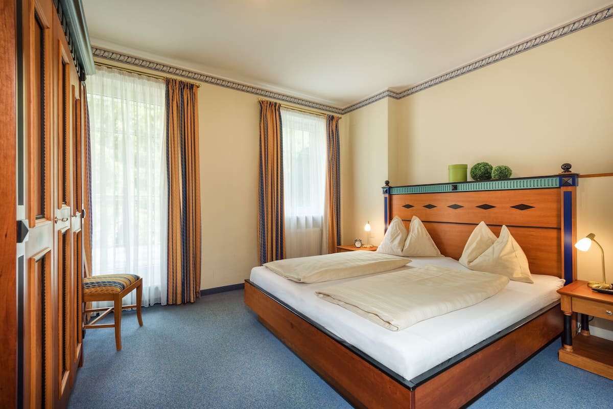 Une chambre d’hôtel avec un grand lit, une tête de lit en bois, une table de chevet avec une lampe, une chaise et une fenêtre avec des rideaux.
