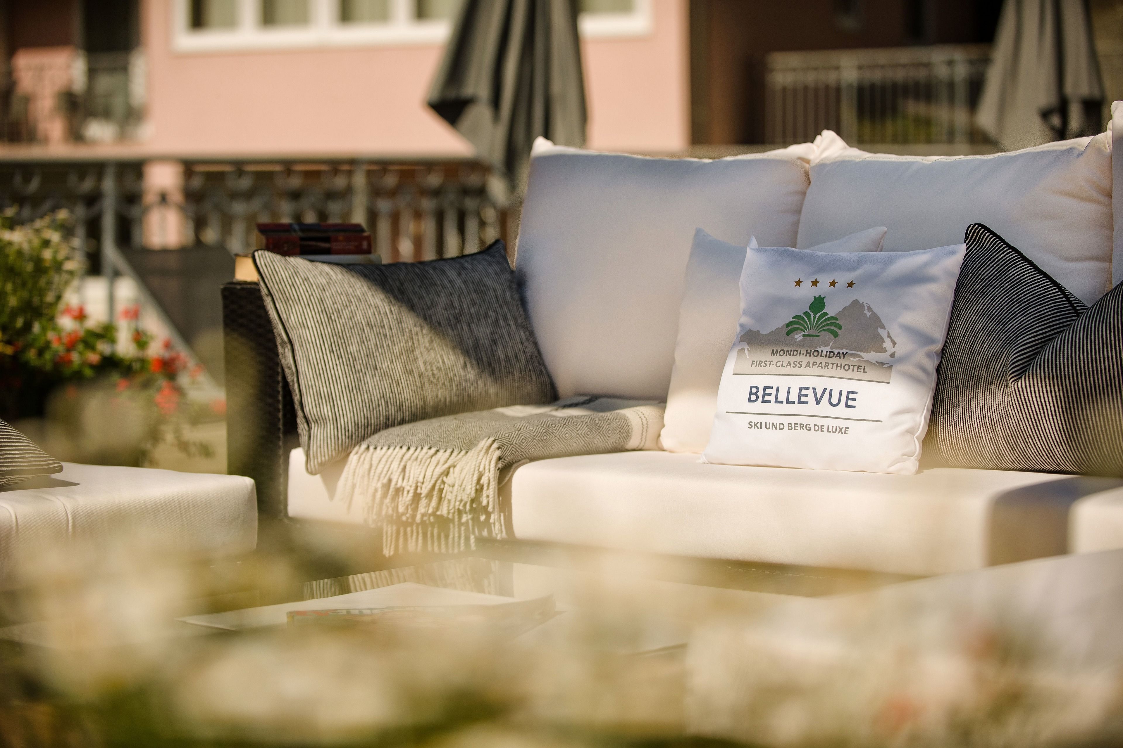 Een witte buitenbank met een plaid en kussens, waaronder een kussen met het merk 'BELLEVUE', in een zonnige omgeving met een gebouw op de achtergrond.