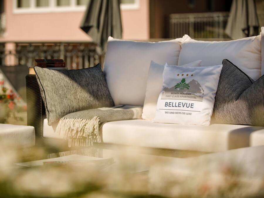 Un sofá blanco para exterior con una manta y almohadones, incluyendo un almohadón de la marca 'BELLEVUE', en un entorno exterior soleado con un edificio al fondo.