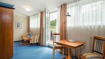 A hotel room with a blue carpet, wooden furniture, a table with a bowl, and a view of greenery through the window.