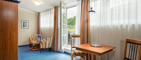 A hotel room with a blue carpet, wooden furniture, a table with a bowl, and a view of greenery through the window.