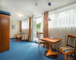 A hotel room with a blue carpet, wooden furniture, a table with a bowl, and a view of greenery through the window.