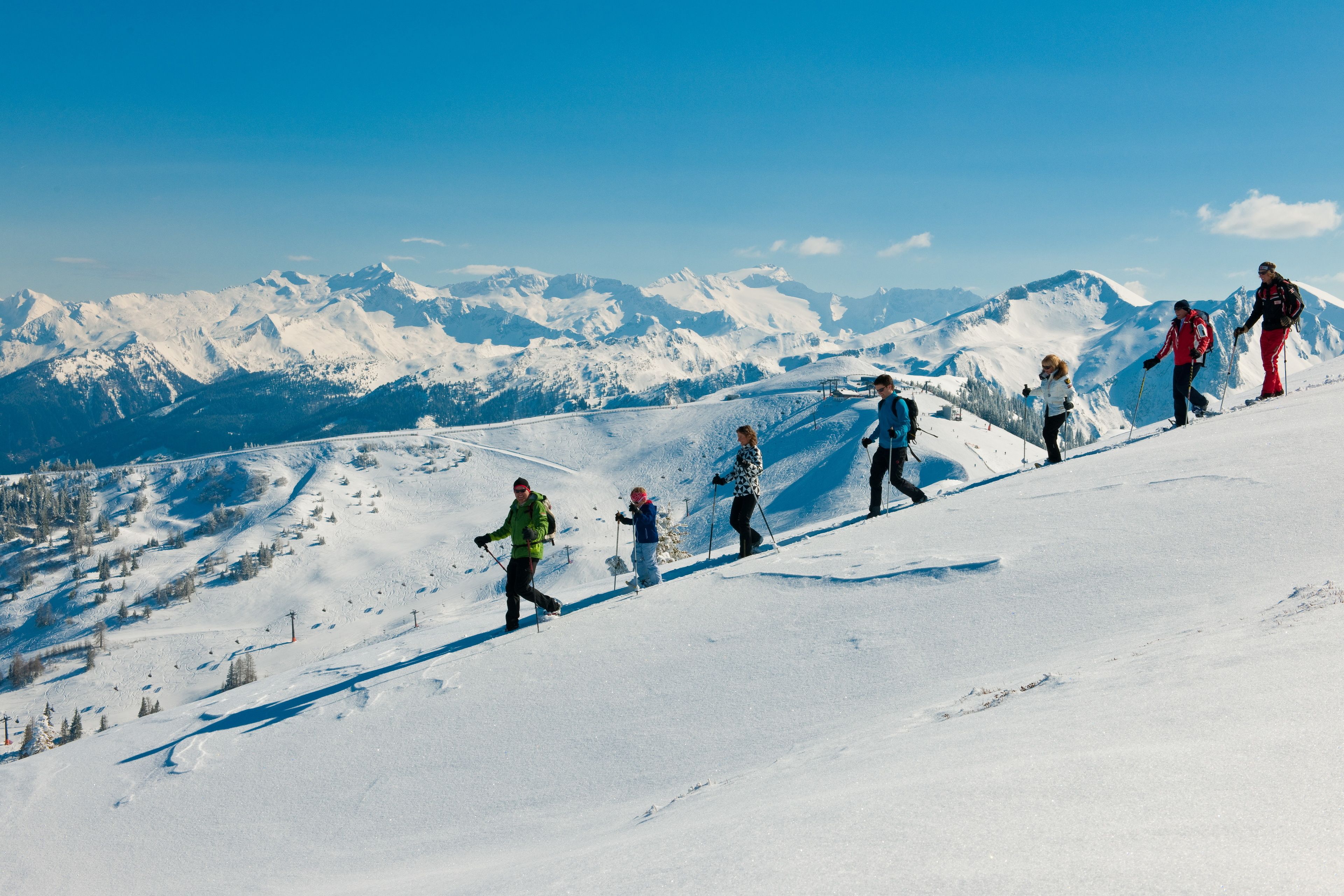 Un grupo de personas caminando cuesta arriba por una ladera nevada, con un cielo azul despejado y picos nevados al fondo.