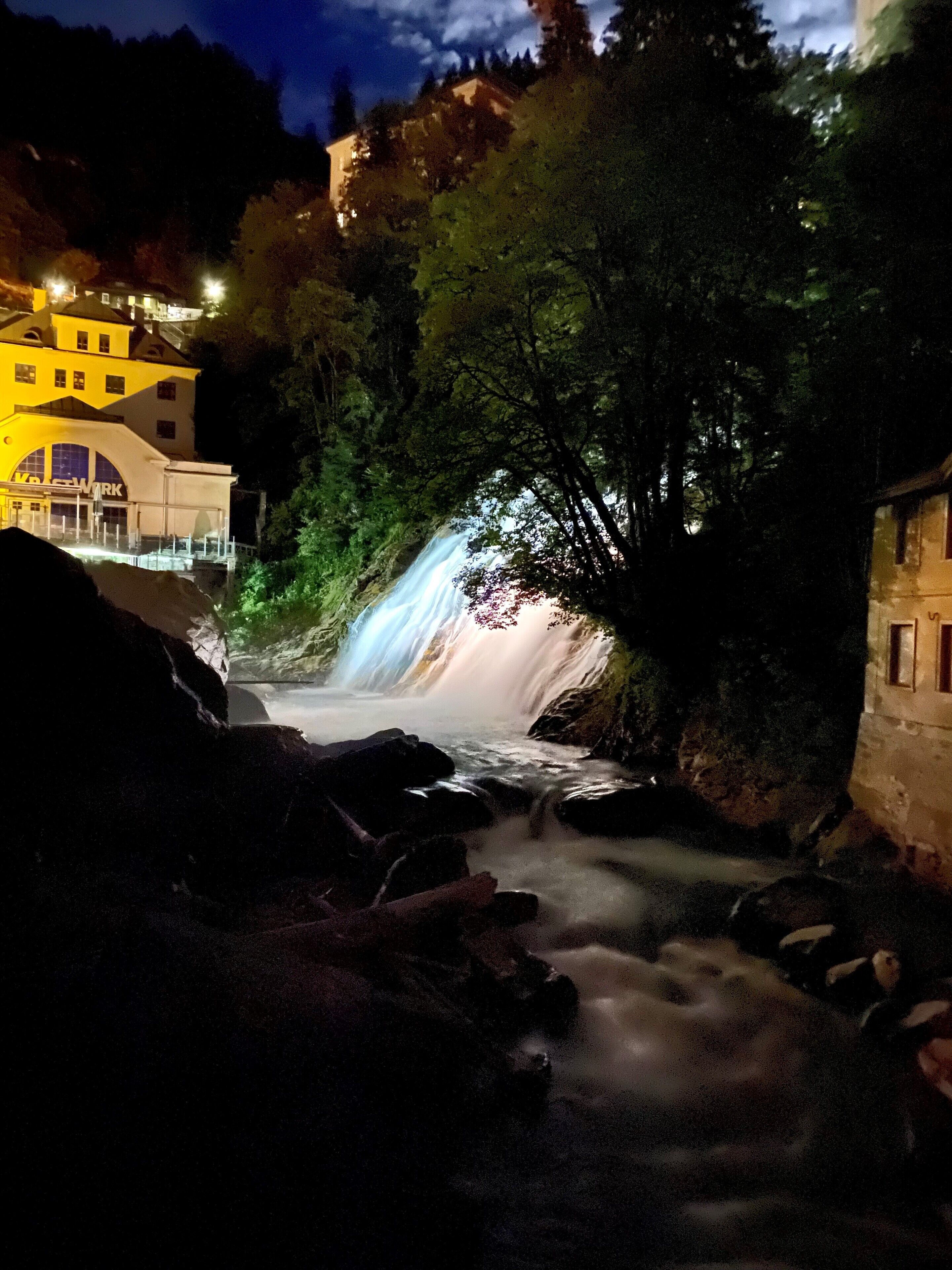 Ein Wasserfall in einer natürlichen Umgebung, im Hintergrund ein Gebäude.