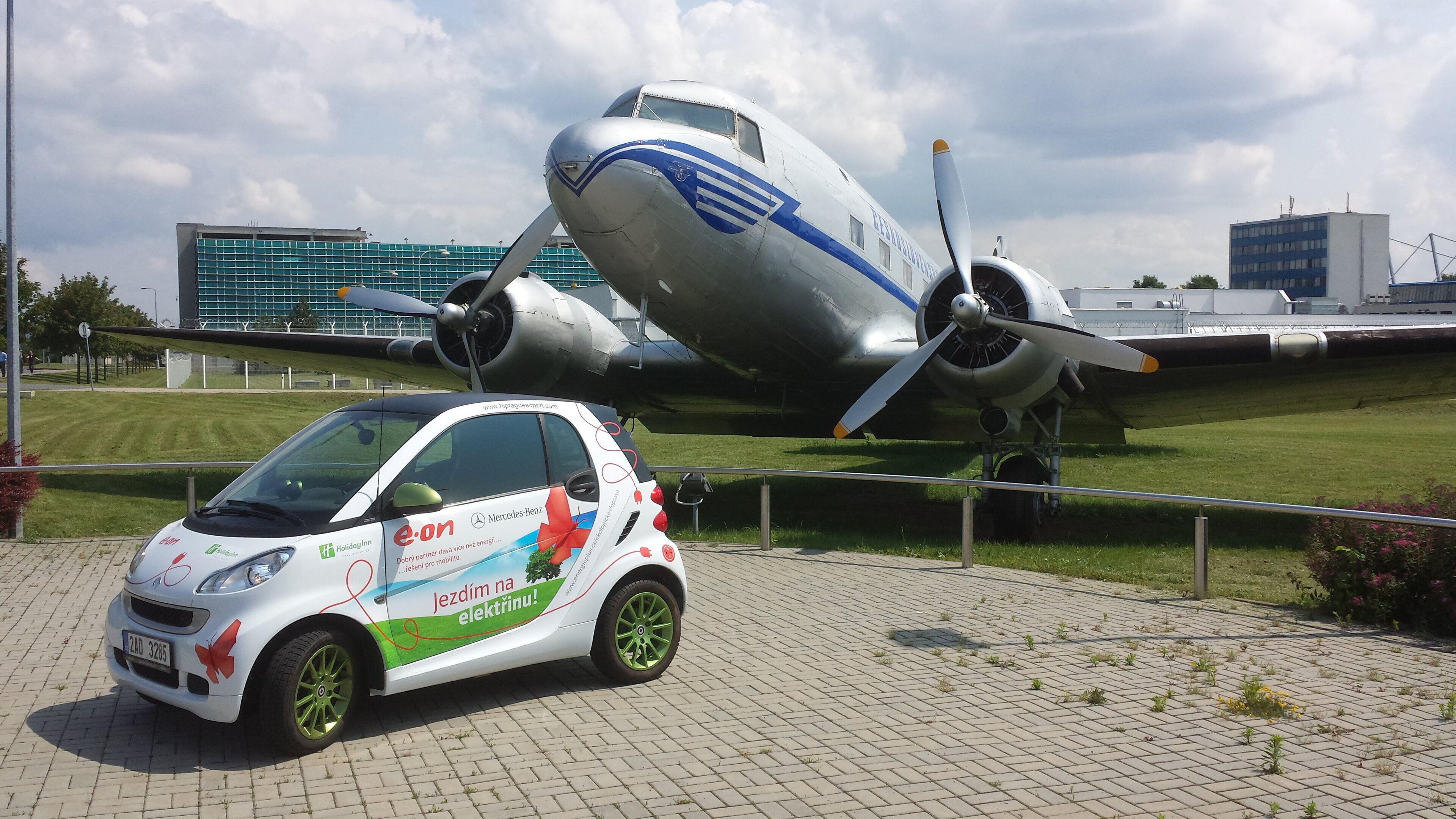 Een witte auto met groene velgen geparkeerd voor een groot vliegtuig.