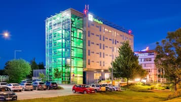 Un hotel Holiday Inn con fachada de vidrio moderna, un estacionamiento con varios coches y un área de césped bien cuidada.