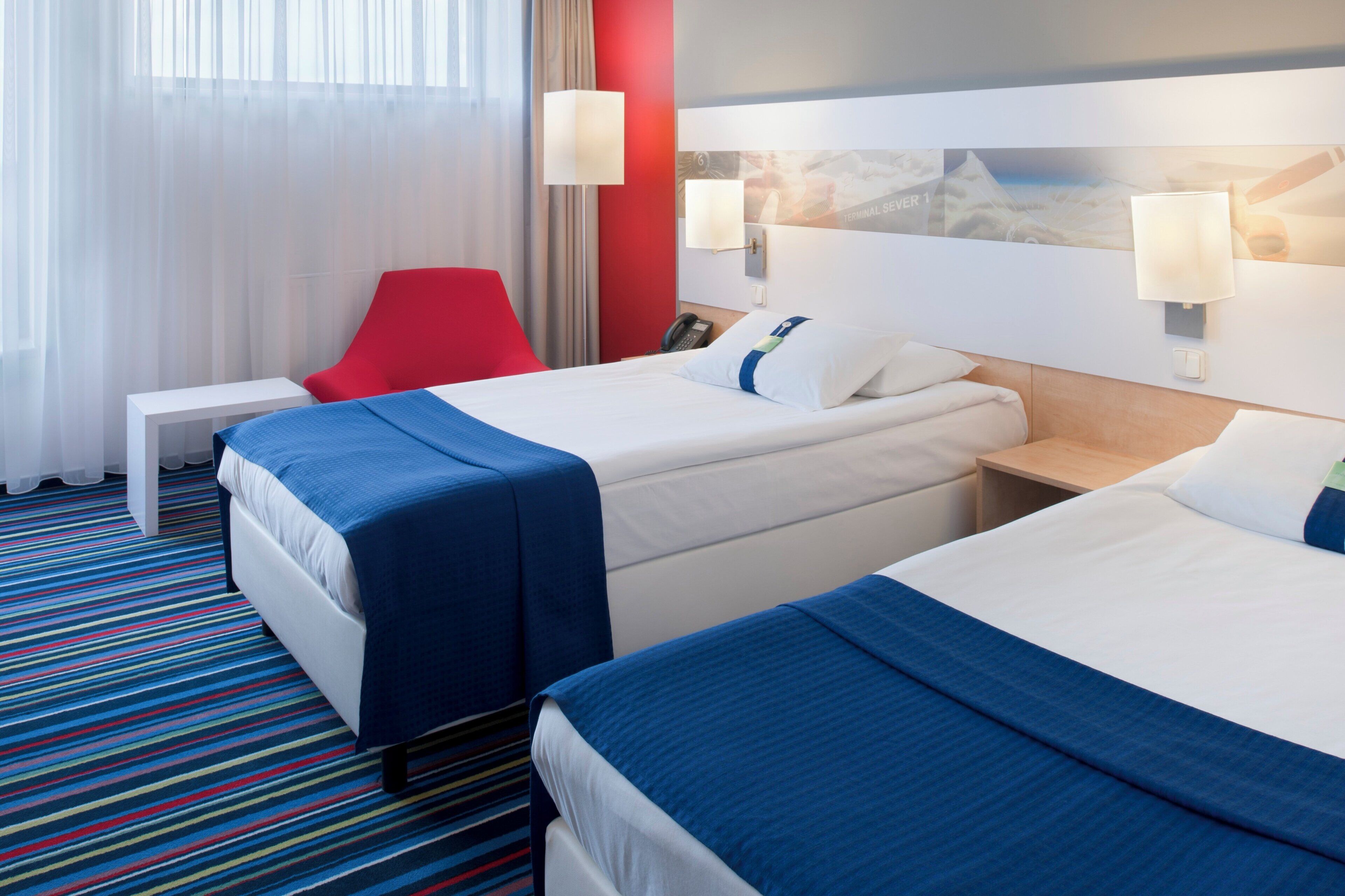 Una habitación de hotel con dos camas, una silla roja, una mesita pequeña y una mesita de noche.
