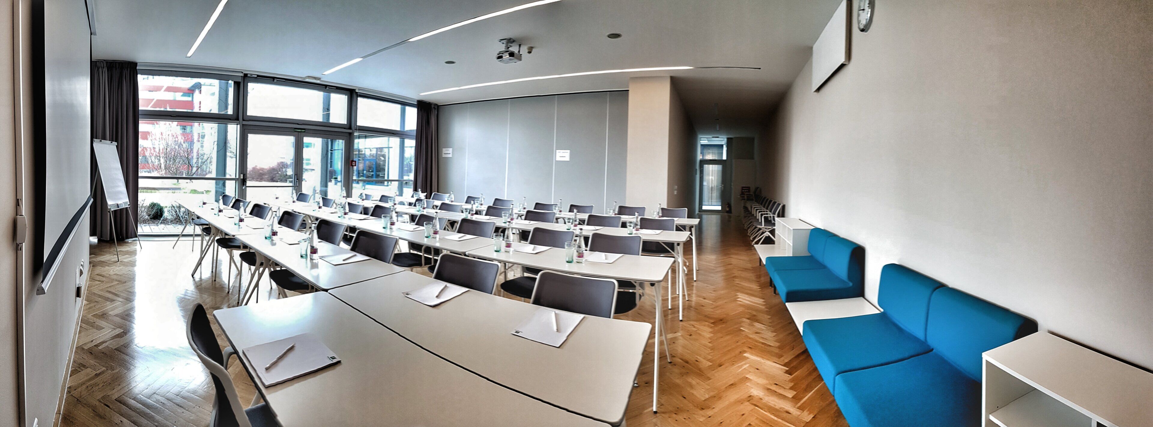 Une salle de conférence dotée de plusieurs tables et chaises, de grandes fenêtres et d’un espace détente aux couleurs bleues.