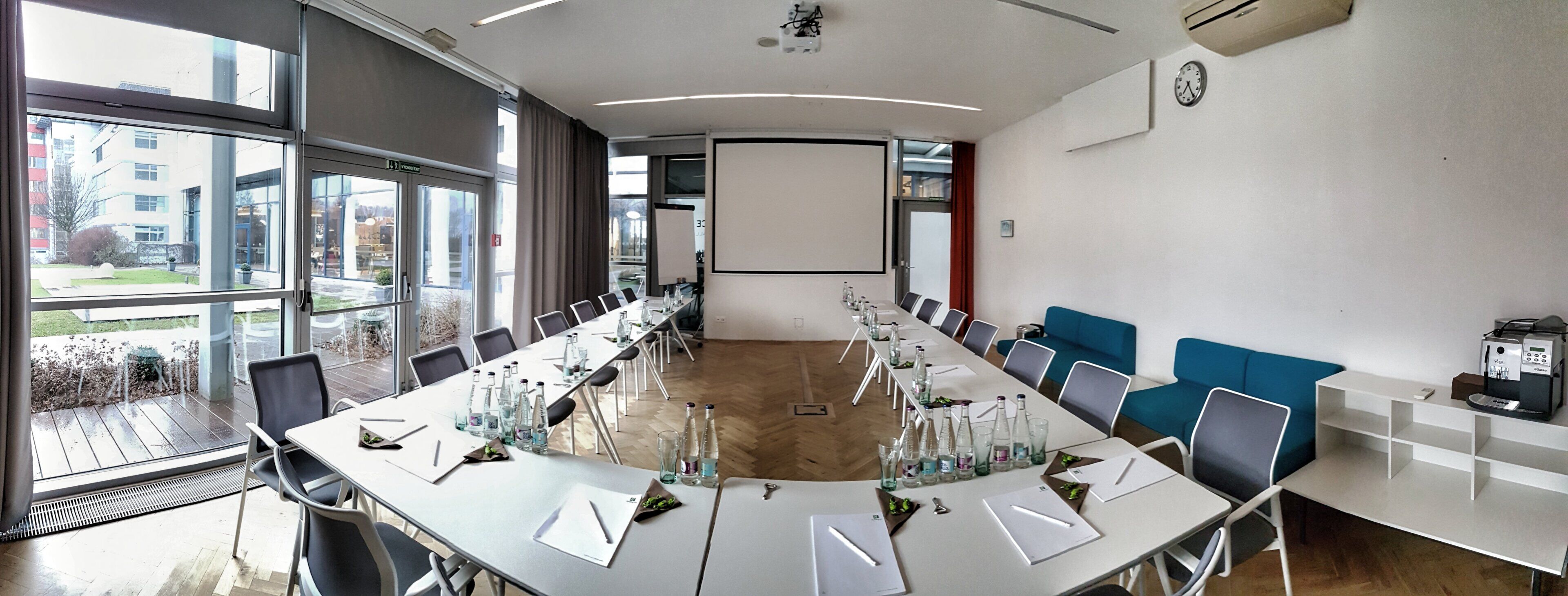 Un salón de conferencias con una larga mesa, sillas, una pantalla de proyector y un reloj en la pared.