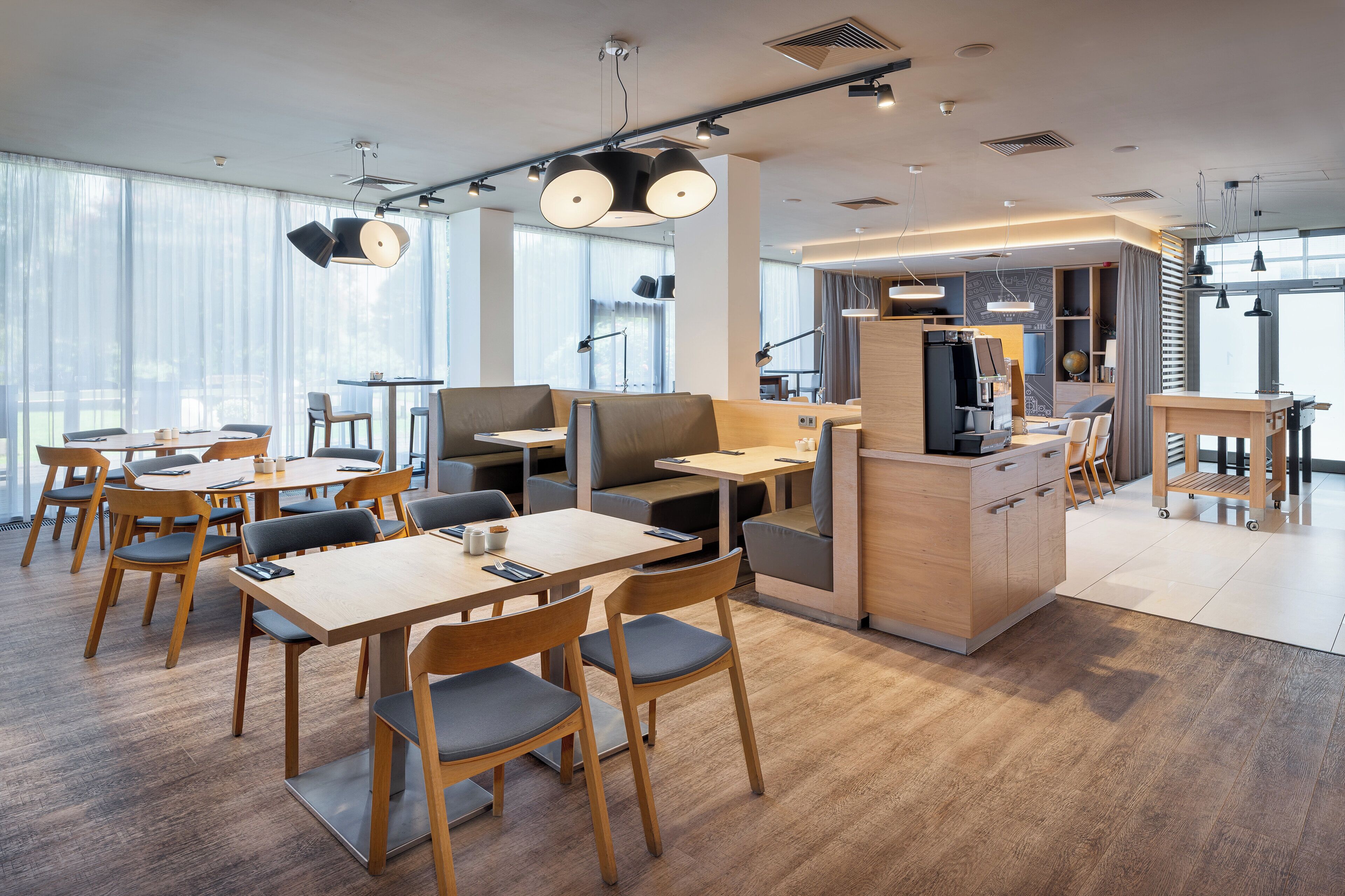 A modern cafe with wooden tables and chairs, a counter with a cash register, and large windows allowing natural light.