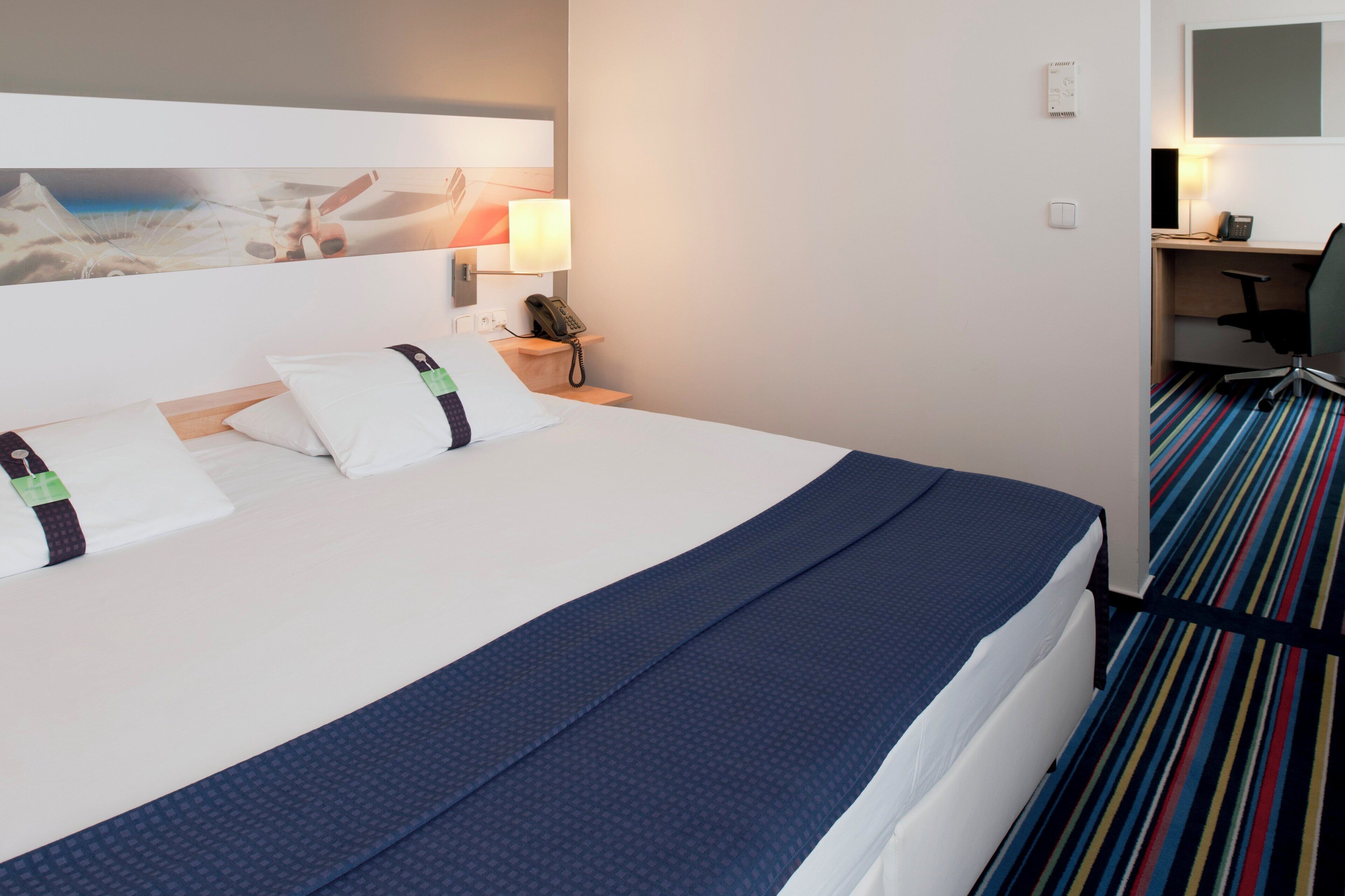 A hotel room with a bed, pillows, a desk with a phone, and a chair.