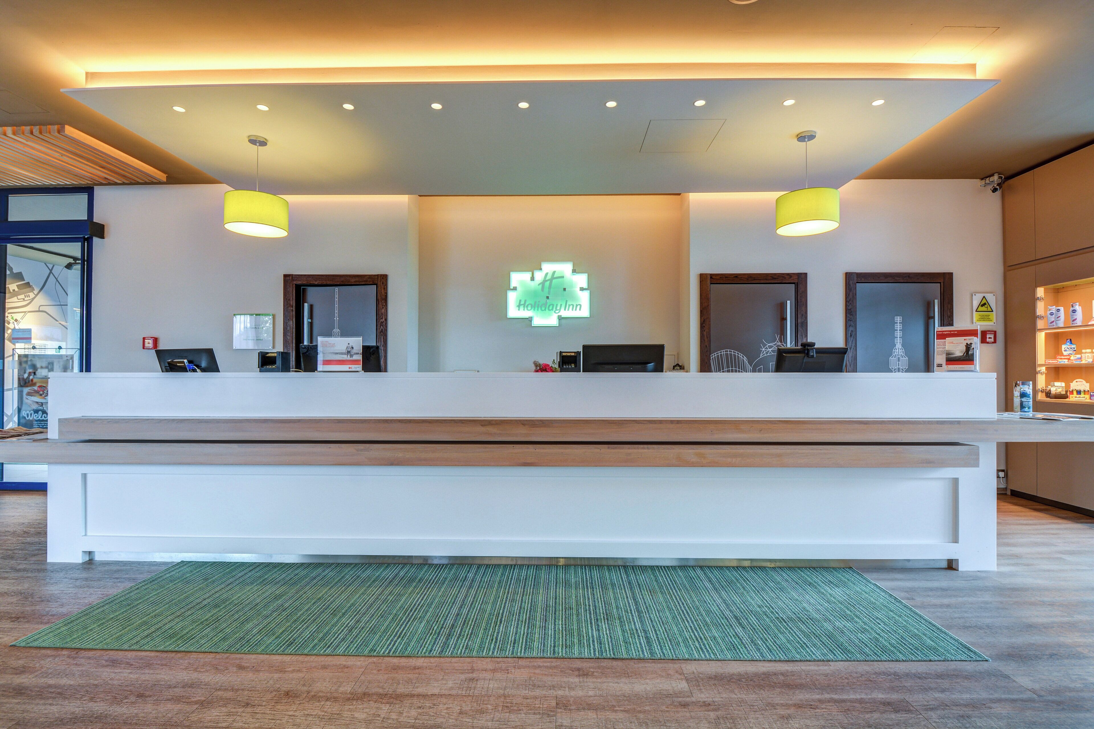 Zona de recepción de un hotel con mostrador, dos lámparas colgantes, alfombra verde y un cartel con el nombre del hotel.