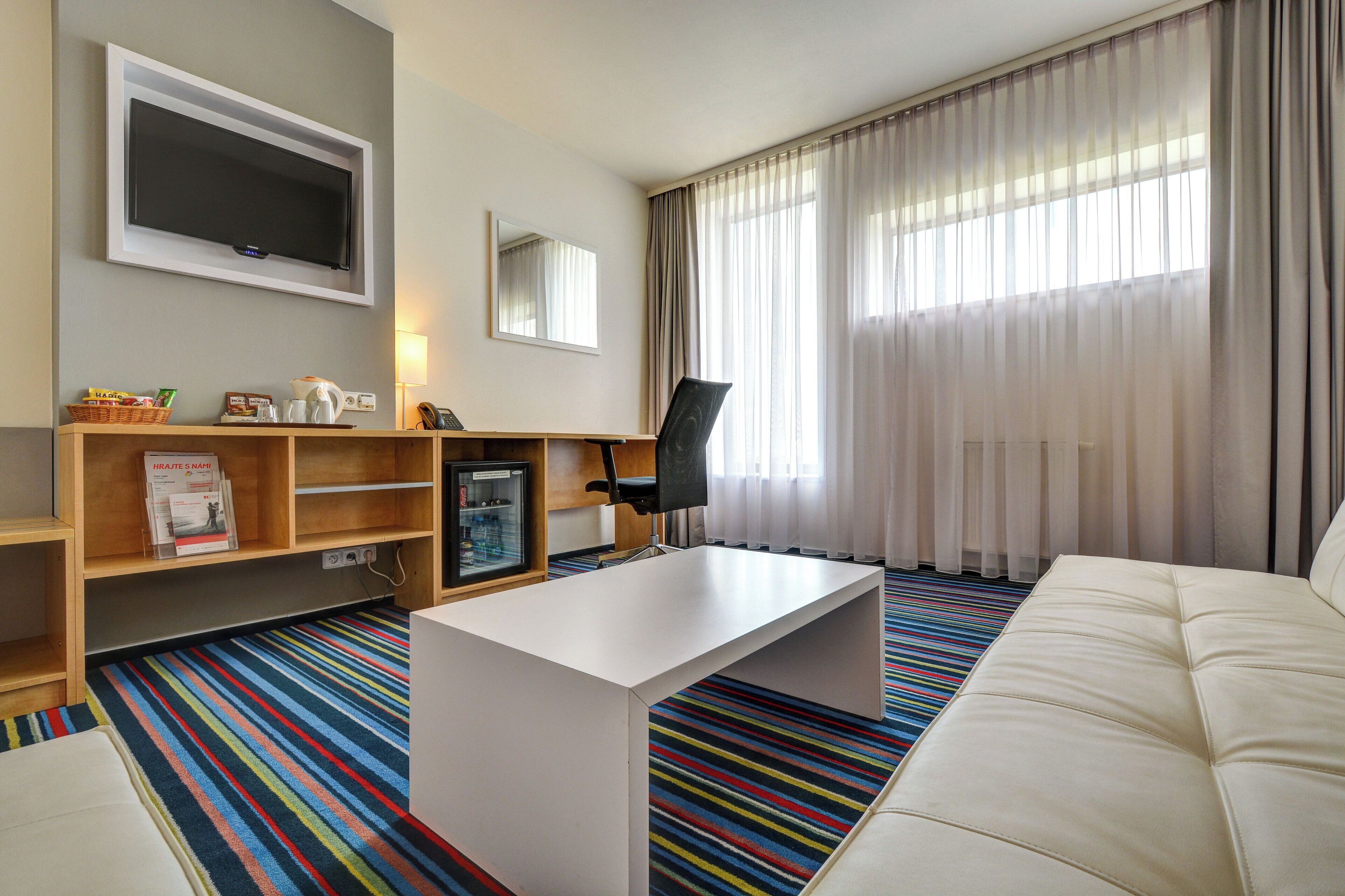 Habitación de hotel con televisión de pantalla plana, mesa de centro, sofá, estantería, refrigerador y ventana con cortinas.