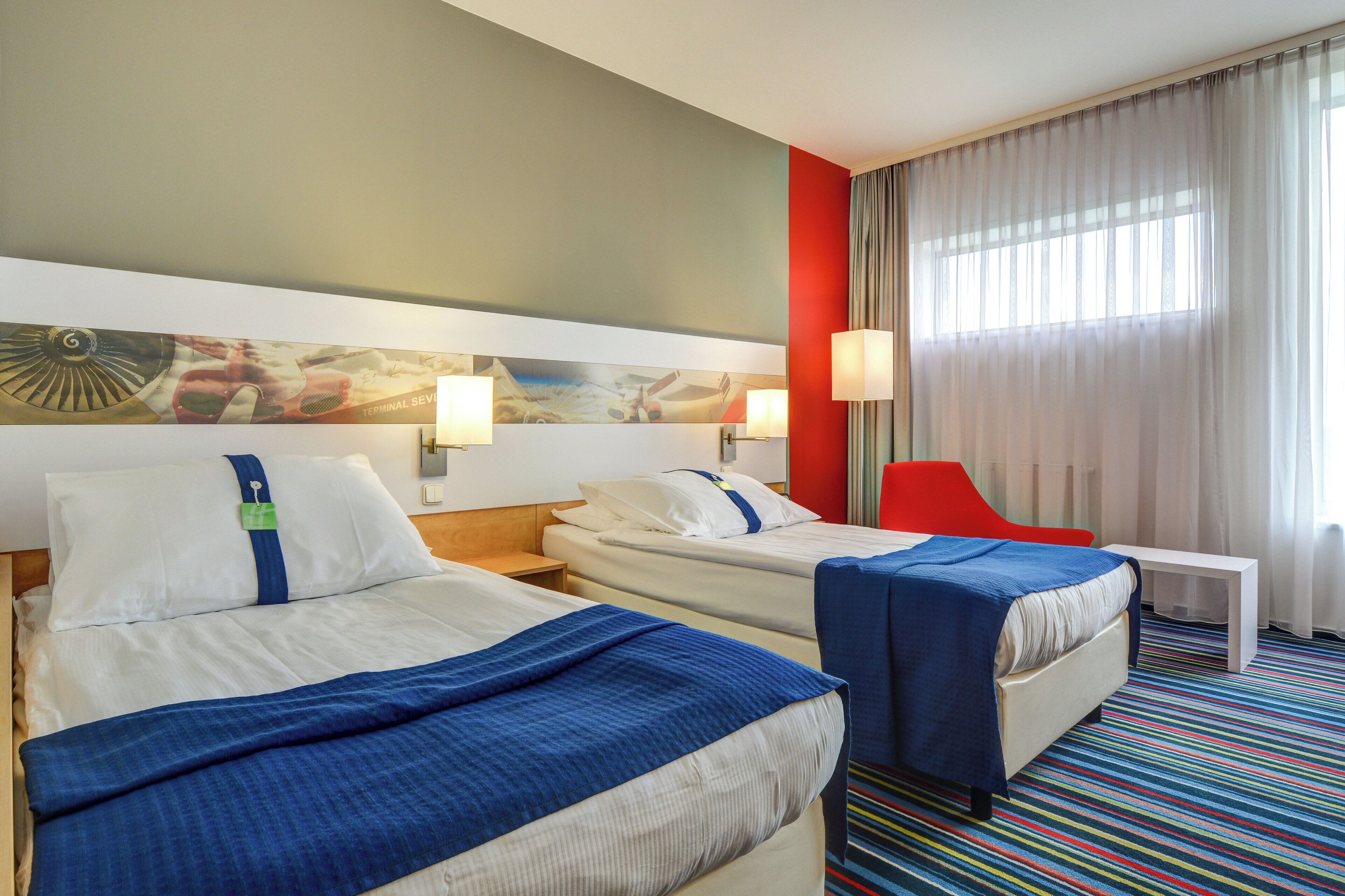 A hotel room with two beds, a red chair, a small table, and a colorful striped carpet.