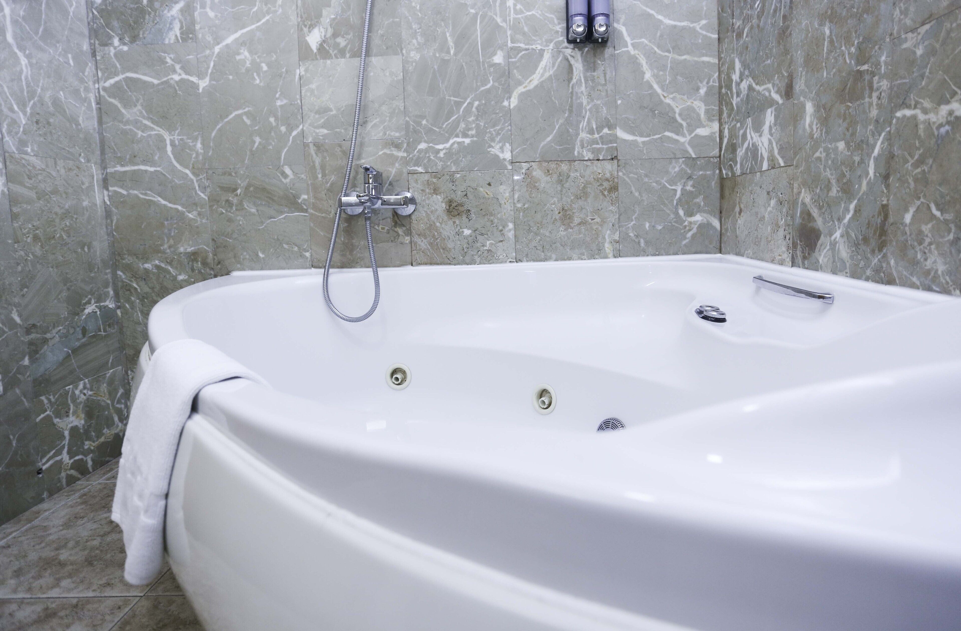 A white bathtub with a shower head and controls, set against a marble wall.