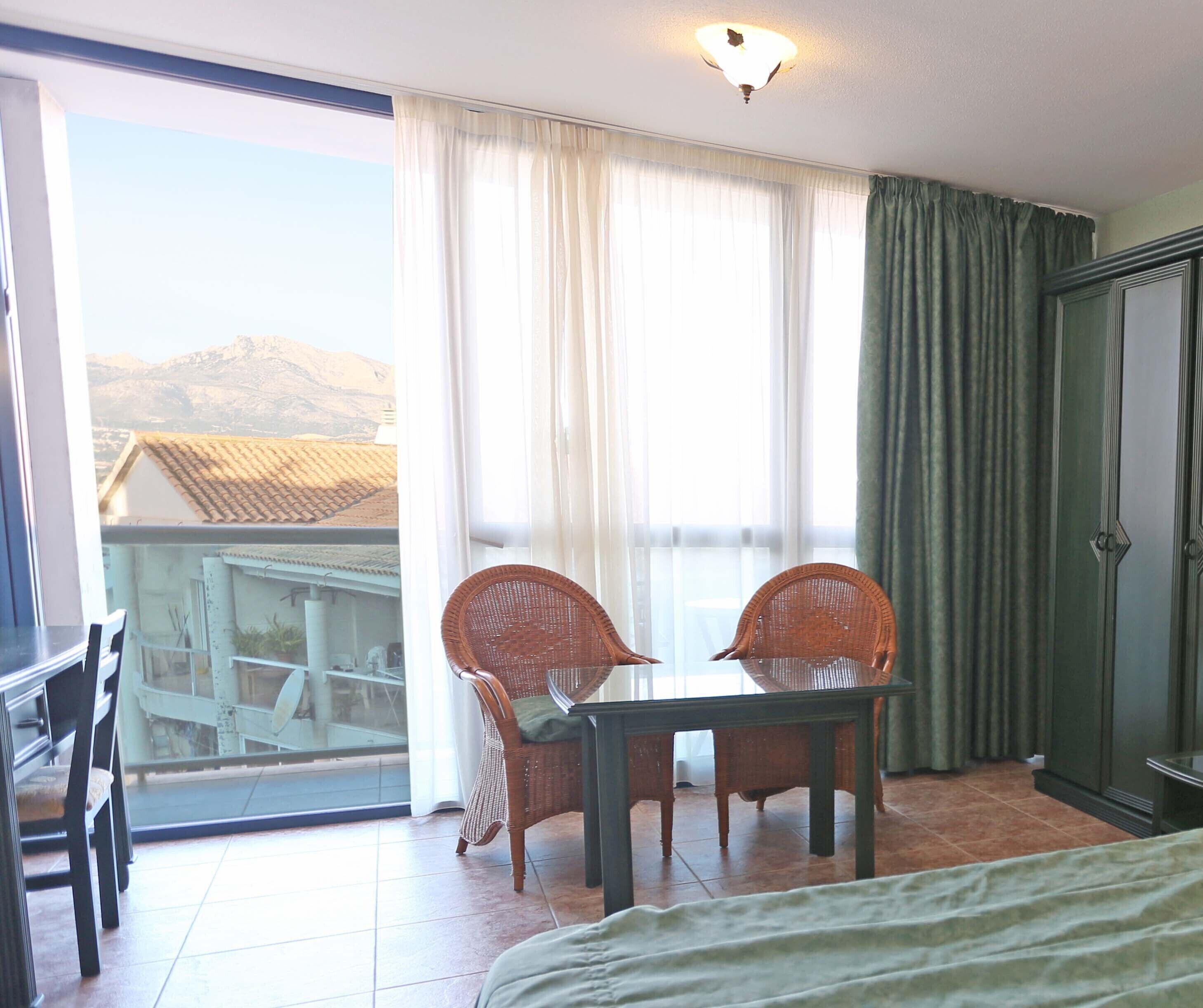 A room with a balcony, a dining table with chairs, a bed, and a wardrobe.