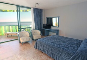 Habitación de hotel con cama, escritorio con televisor, dos sillas, ventana grande con vista al mar y balcón.