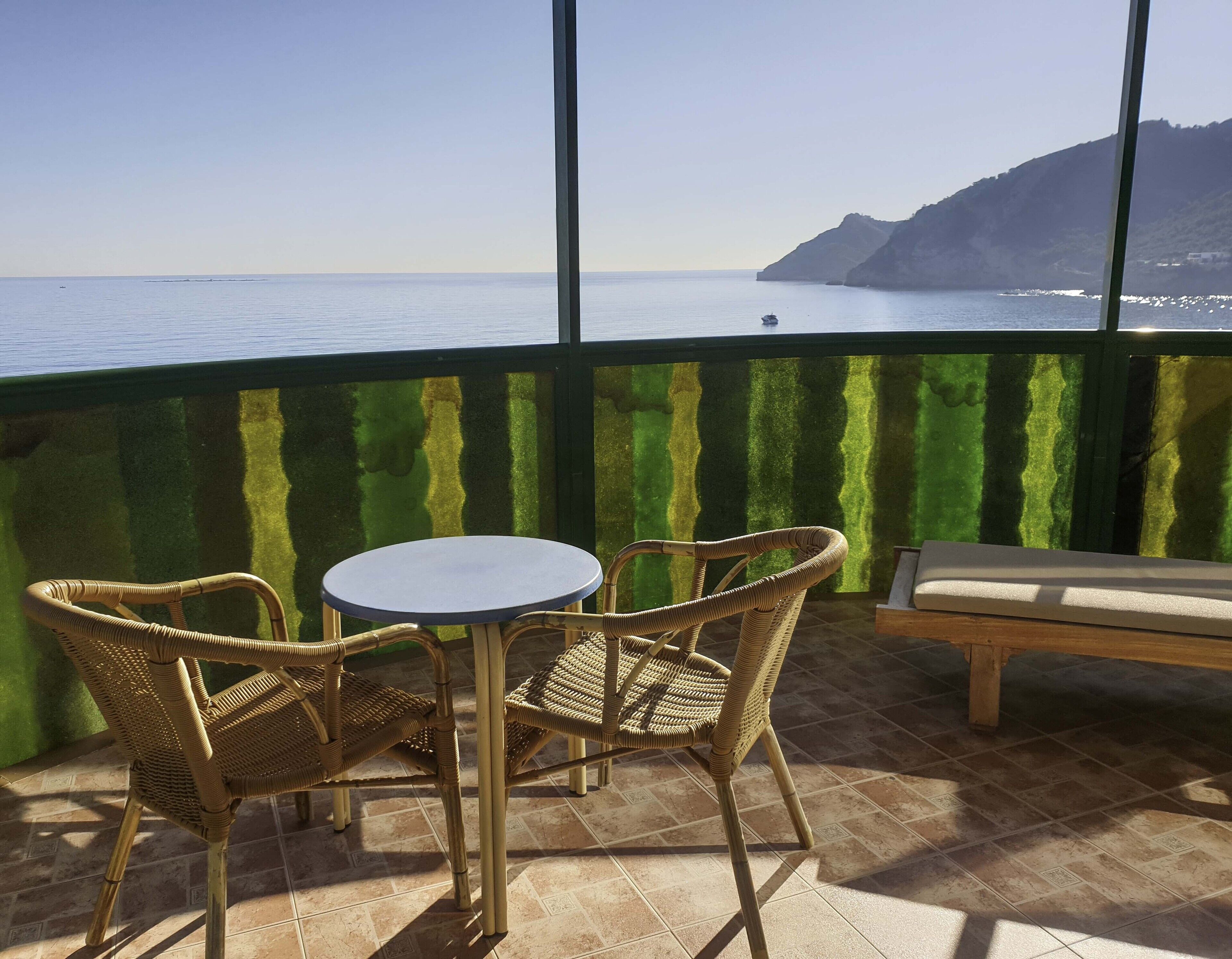 A balcony with wicker chairs and a table, overlooking a calm sea and a mountainous landscape.