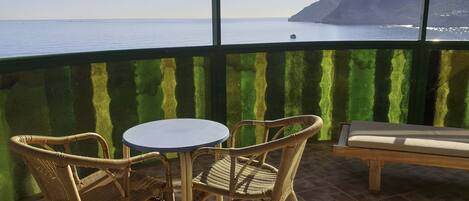 A balcony with wicker chairs and a table, overlooking a calm sea and a mountainous landscape.