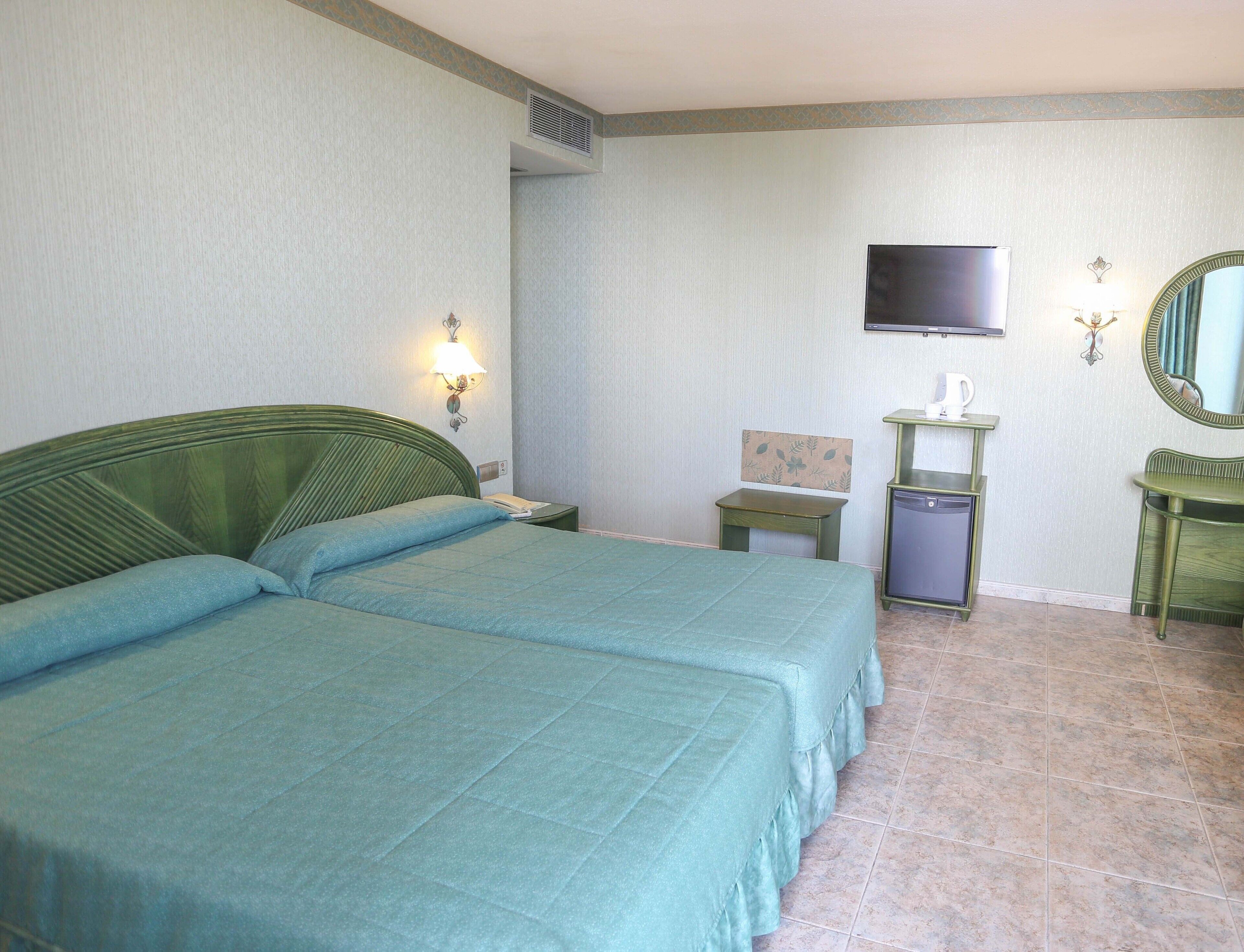A hotel room with two beds, a television, a small table, a mirror, and a wall-mounted heater.