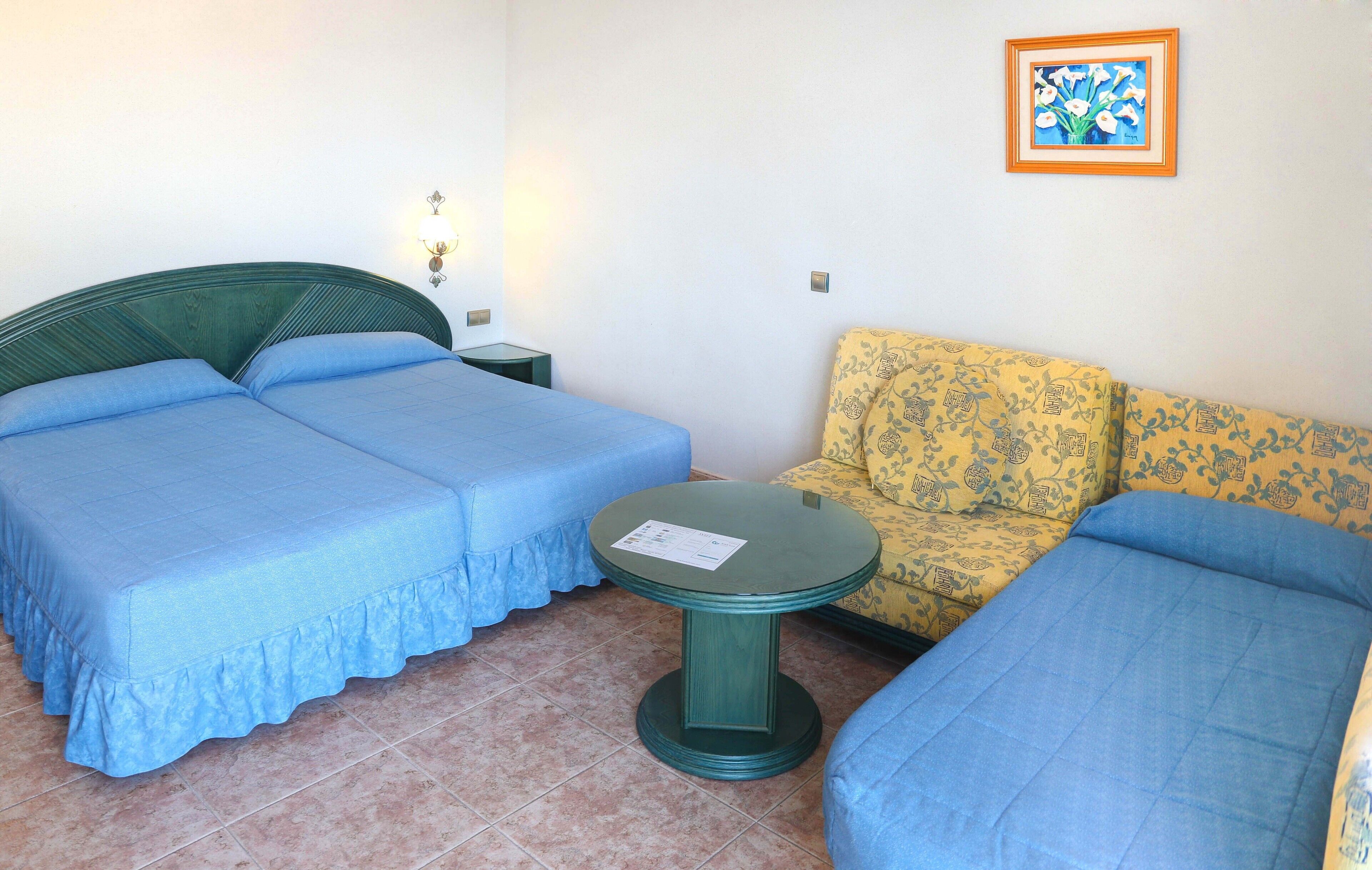 A room with two single beds, a small round table, a sofa, and a wall-mounted picture.