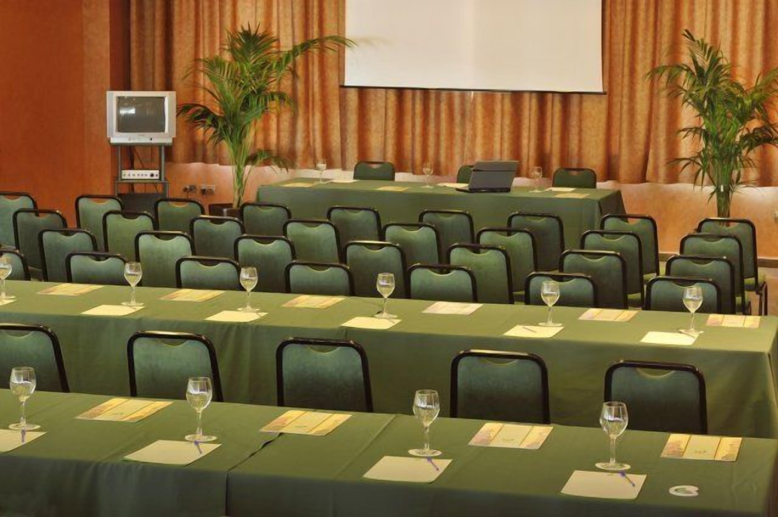 Une salle de conférence avec des rangées de chaises vertes, une grande table sur laquelle se trouve un ordinateur portable et un écran de projection.