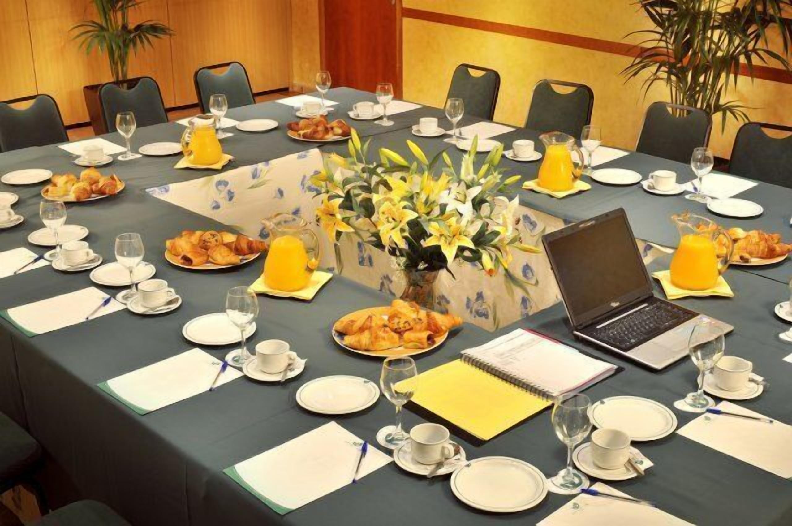 Une salle de conférence avec une longue table dressée pour le petit-déjeuner, comprenant des assiettes, des tasses et un ordinateur portable.