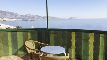 Un balcón con una silla de mimbre y una mesa pequeña, con vista al mar, montañas y cielo despejado.
